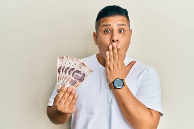 Genç Latin adam 500 Meksika pezosu tutuyor. Ağzını eliyle kapatıyor. Şaşırmış ve hata yapmaktan korkuyor. şaşırmış ifade 
