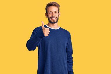 Sakallı, sıradan süveter giyen yakışıklı sarışın el hareketleriyle mutlu mesut el hareketi yapıyor. Kameraya bakıp başarıyı gösteren ifade. 