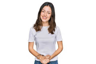 Genç, güzel bir kadın, elleri çapraz ve günlük beyaz tişörtler giyiyor. Rahatlamış ve neşeli gülümsüyor. Başarı ve iyimserlik 