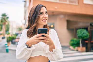 Genç İspanyol kadın şehirde akıllı telefon kullanarak mutlu bir şekilde gülümsüyor..