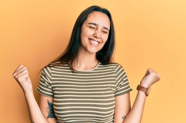 Genç İspanyol kadın rahat çizgili tişört giyiyor. Çok mutlu ve heyecanlı. Kolları havada, gülümseyen ve başarı için çığlık atan kazanan bir jest yapıyor. kutlama konsepti. 