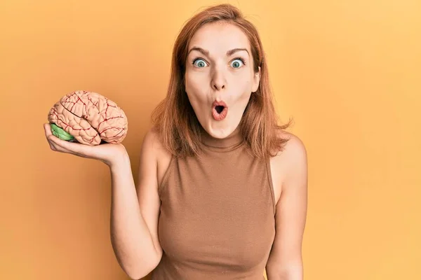 Young caucasian woman holding brain scared and amazed with open mouth for surprise, disbelief face