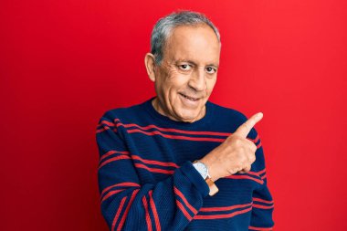 Kıdemli İspanyol adam, günlük kıyafetler giyiyor. Gülümsüyor, eli ve parmağı yana doğru uzanıyor. 