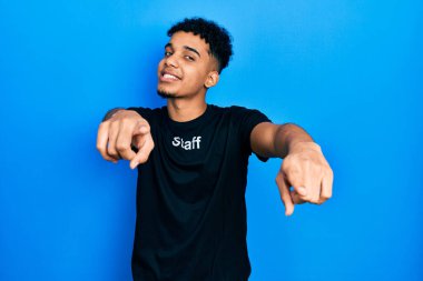 Genç, Afro-Amerikalı, personel tişörtü giymiş. Kamerayı ve seni işaret eden parmakları olan, neşeli ve pozitif gülümseyen adam. 