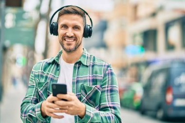 Genç beyaz adam şehirde akıllı telefon ve kulaklık kullanarak mutlu bir şekilde gülümsüyor..