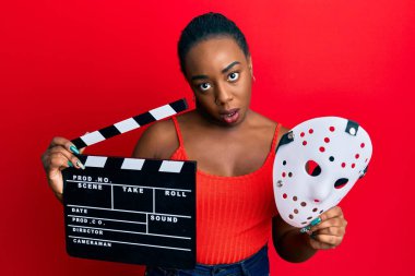 Genç Afro-Amerikalı kadın elinde video film panosu ve korku maskesiyle şaşkın, şüpheci ve iğneleyici bakıyor, ağzı açık kalınca şaşırıyor. 