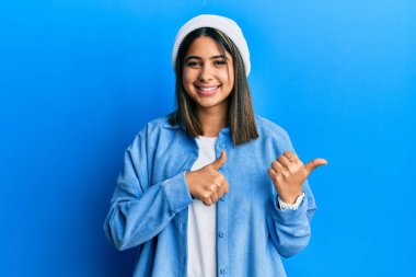 Arkasını gösteren sevimli yün şapkalı genç Latin kadın. Baş parmakları havada, kendinden emin gülümsüyor. 