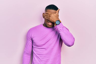 Sıradan pembe kazak giyen genç siyahi bir adam. Gülümsüyor ve elleri yüzlerinde, şaşırtmak için gözleri örtüyor. kör kavram. 