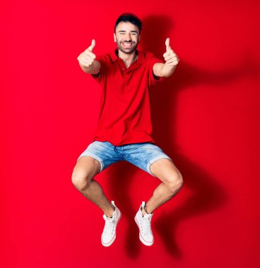 Şapkalı genç teslimatçı mutlu gülümsüyor. Yüzünde tebessümle zıplıyor. Tamam işareti yapıyor. Kırmızı arka plan üzerinde başparmaklar yukarı doğru.