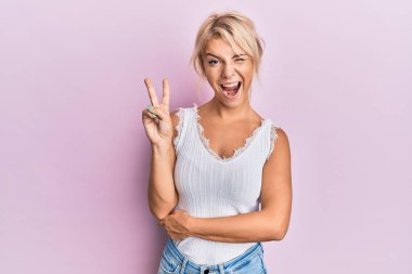 Sıradan kıyafetler giyen, gülümseyen, kameraya göz kırpan, zafer işareti yapan genç sarışın kız. İki numara.. 