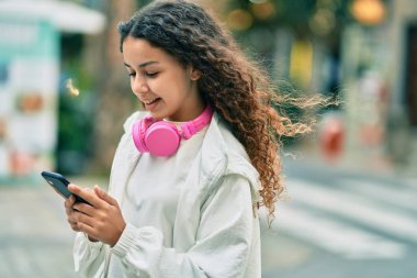 Güzel İspanyol kız şehirde akıllı telefon ve kulaklık kullanarak mutlu bir şekilde gülümsüyor..
