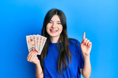 Young beautiful caucasian girl holding 10 united kingdom pounds banknotes smiling happy pointing with hand and finger to the side 