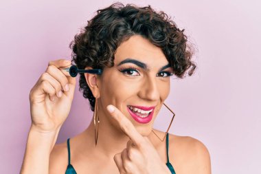 Kirpik rimel tutan genç adam gülümseyerek el ve parmağıyla işaret ediyor. 