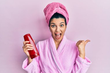 Duş bornozu giymiş, elinde şampuan tutan genç İspanyol kadın başparmağını kaldırıyor ve ağzı açık gülümsüyor. 