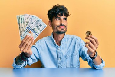 Elinde dolar ve bitcoin tutan genç İspanyol adam gülümsüyor ve yan tarafa bakıyor ve düşünüyor. 