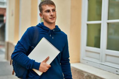 Üniversitede elinde defterle gülümseyen sarışın genç bir öğrenci..