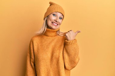 Orta yaşlı, beyaz kadın yün kazak ve şapkalı, gülen yüzlü ve baş parmağıyla yüzünü gösteren.. 