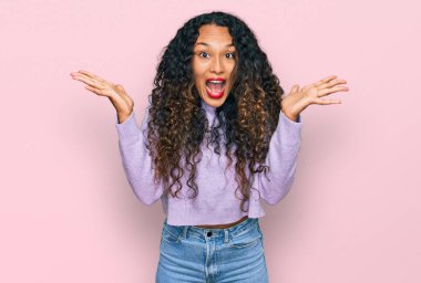 Young hispanic woman with curly hair wearing casual winter sweater celebrating victory with happy smile and winner expression with raised hands 