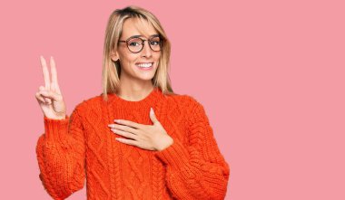 Güzel sarışın kadın, gündelik kıyafetler ve gözlük takıyor, gülümsüyor, yemin ediyor, el ve göğsü havada küfrediyor. 