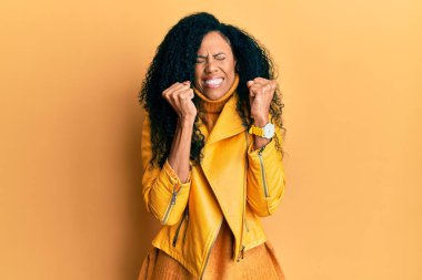 Orta yaşlı Afro-Amerikalı kadın yün kazak ve deri ceket giyiyor. Kolları havada ve gözleri kapalı zaferi kutlarken gülümsüyor. Kazanan konsept. 