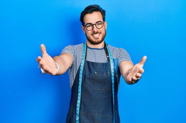 Handsome caucasian man with beard dressmaker designer wearing atelier apron looking at the camera smiling with open arms for hug. cheerful expression embracing happiness. 