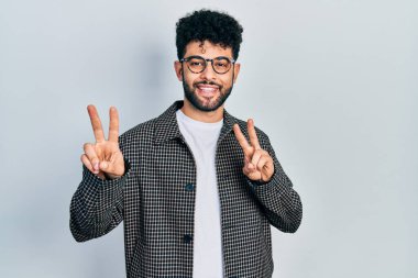 Sakallı, gözlüklü, gülümseyen, parmaklarını zafer işareti yaparken gösteren genç bir Arap. İki numara.. 