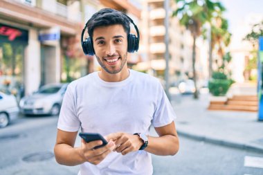 Genç Latin adam şehirde akıllı telefon ve kulaklık kullanarak mutlu bir şekilde gülümsüyor..