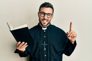 Young hispanic priest man holding bible with finger up winking looking at the camera with sexy expression, cheerful and happy face. 