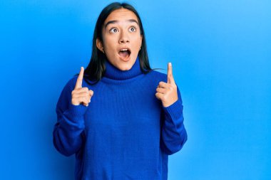 Günlük kışlık süveter giyen genç Asyalı kadın şaşırmış ve şaşırmış bir şekilde yukarı baktı ve parmaklarıyla işaret ederek kollarını kaldırdı.. 