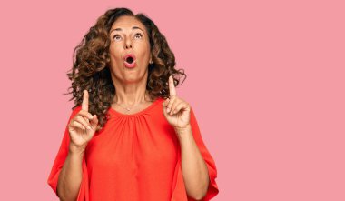 Orta yaşlı İspanyol kadın gündelik kıyafetler giyip şaşkın şaşkın yukarı bakıyor ve parmaklarıyla işaret ediyor ve kollarını kaldırıyor.. 