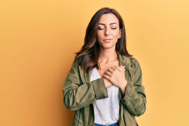 Sarı arka plan üzerine gündelik kıyafetler giyen genç esmer kadın elleri göğsünde, gözleri kapalı ve yüzünde minnet dolu bir jest ile gülümsüyor. sağlık kavramı. 