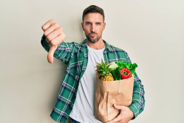 Handsome man with beard holding paper bag with groceries looking unhappy and angry showing rejection and negative with thumbs down gesture. bad expression. 