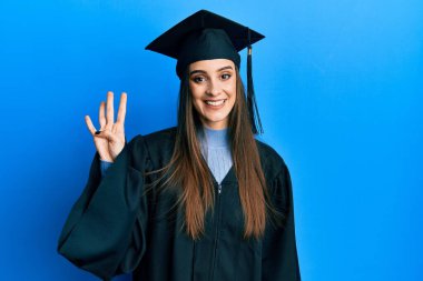 Güzel esmer kadın mezuniyet şapkası ve tören cübbesi takıyor. Dört numaralı parmaklarıyla işaret ediyor ve gülümsüyor. Kendinden emin ve mutlu.. 