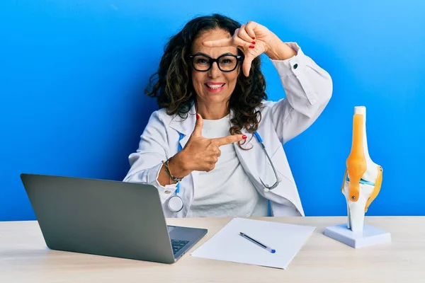 Beautiful middle age woman doctor at orthopedic clinic smiling making frame with hands and fingers with happy face. creativity and photography concept.