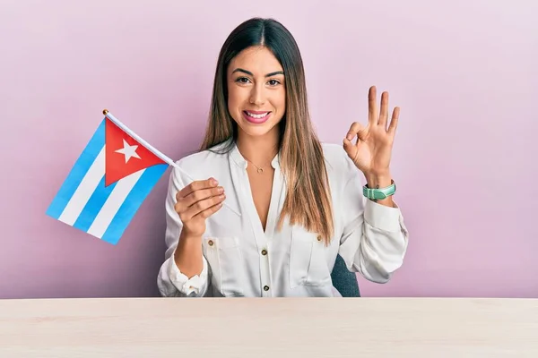 若いですヒスパニック系の女性を保持キューバのフラグは テーブルの上に座って指でOkサインを行います 笑顔優しいジェスチャー優れたシンボル — ストック写真