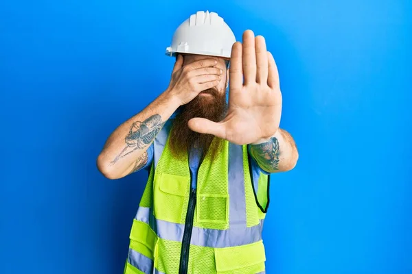 Redhead man with long beard wearing safety helmet and reflective jacket covering eyes with hands and doing stop gesture with sad and fear expression. embarrassed and negative concept.