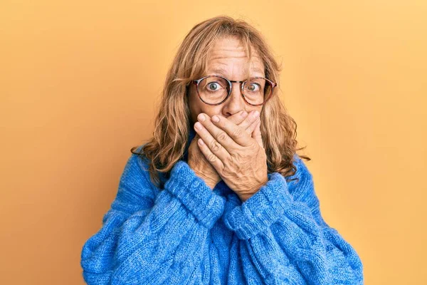 Middle age blonde woman wearing glasses and casual winter sweater shocked covering mouth with hands for mistake. secret concept.