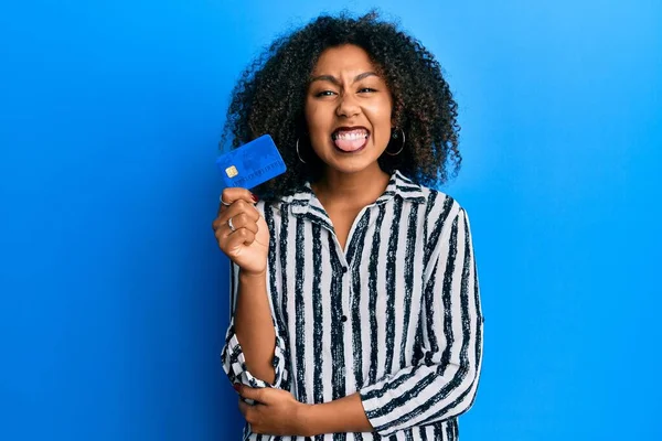 Beautiful african american woman with afro hair holding credit card sticking tongue out happy with funny expression.