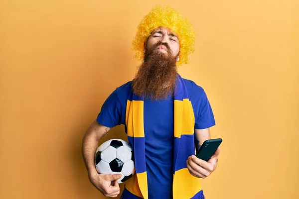 Redhead man with long beard football hooligan cheering game holding smartphone looking at the camera blowing a kiss being lovely and sexy. love expression.