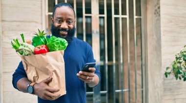 Sakallı Afrikalı Amerikalı adam elinde süpermarketten alınmış kağıt torbalarla akıllı telefon kullanıyor.