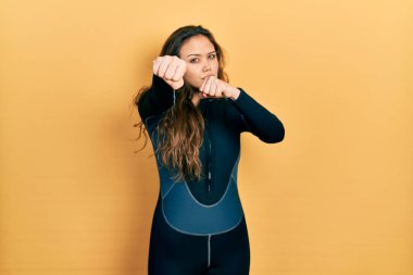 Young hispanic girl wearing diver neoprene uniform punching fist to fight, aggressive and angry attack, threat and violence 