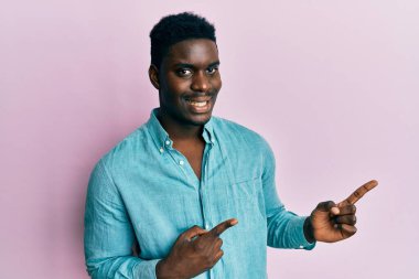 Yakışıklı, sıradan kıyafetler giyen, gülümseyen ve kameraya bakan, iki eli ve parmağı yanda olan siyahi bir adam.. 