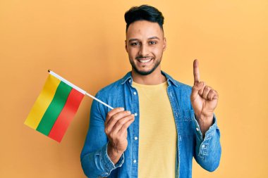 Elinde Litvanya bayrağı olan genç bir Arap adam bir fikir veya soruyla gülümsüyor ve mutlu bir yüzle işaret ediyor. 