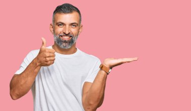 Orta yaşlı beyaz tişört giyen yakışıklı bir adam avuç içini gösteriyor ve baş parmaklarıyla iyi bir hareket yapıyor, mutlu ve neşeli gülümsüyor. 