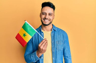 Genç Arap adam senegal bayrağını tutuyor. Pozitif ve mutlu görünüyor. Duruyor ve gülümsüyor. 