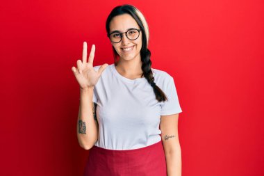 Genç İspanyol kadın profesyonel garson önlüğü giyiyor. Üç numaralı parmaklarıyla işaret ediyor. Kendinden emin ve mutlu bir şekilde gülümsüyor.. 