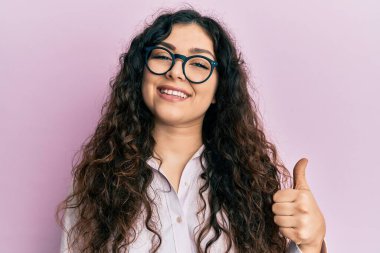 Young brunette woman with curly hair wearing casual clothes and glasses doing happy thumbs up gesture with hand. approving expression looking at the camera showing success. 