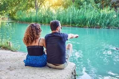 Sıradan kıyafetler giyen orta yaşlı bir çift. Nehrin gerisinde oturuyor, bakıyor ve parmağıyla nehri işaret ediyor..