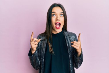 Parti ceketi giyen genç İspanyol kız şaşırmış ve şaşırmış bir şekilde yukarı bakıyor ve parmaklarıyla işaret ediyor ve kollarını kaldırıyor.. 