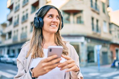 Beyaz kadın şehirde akıllı telefon ve kulaklık kullanarak mutlu bir şekilde gülümsüyor..